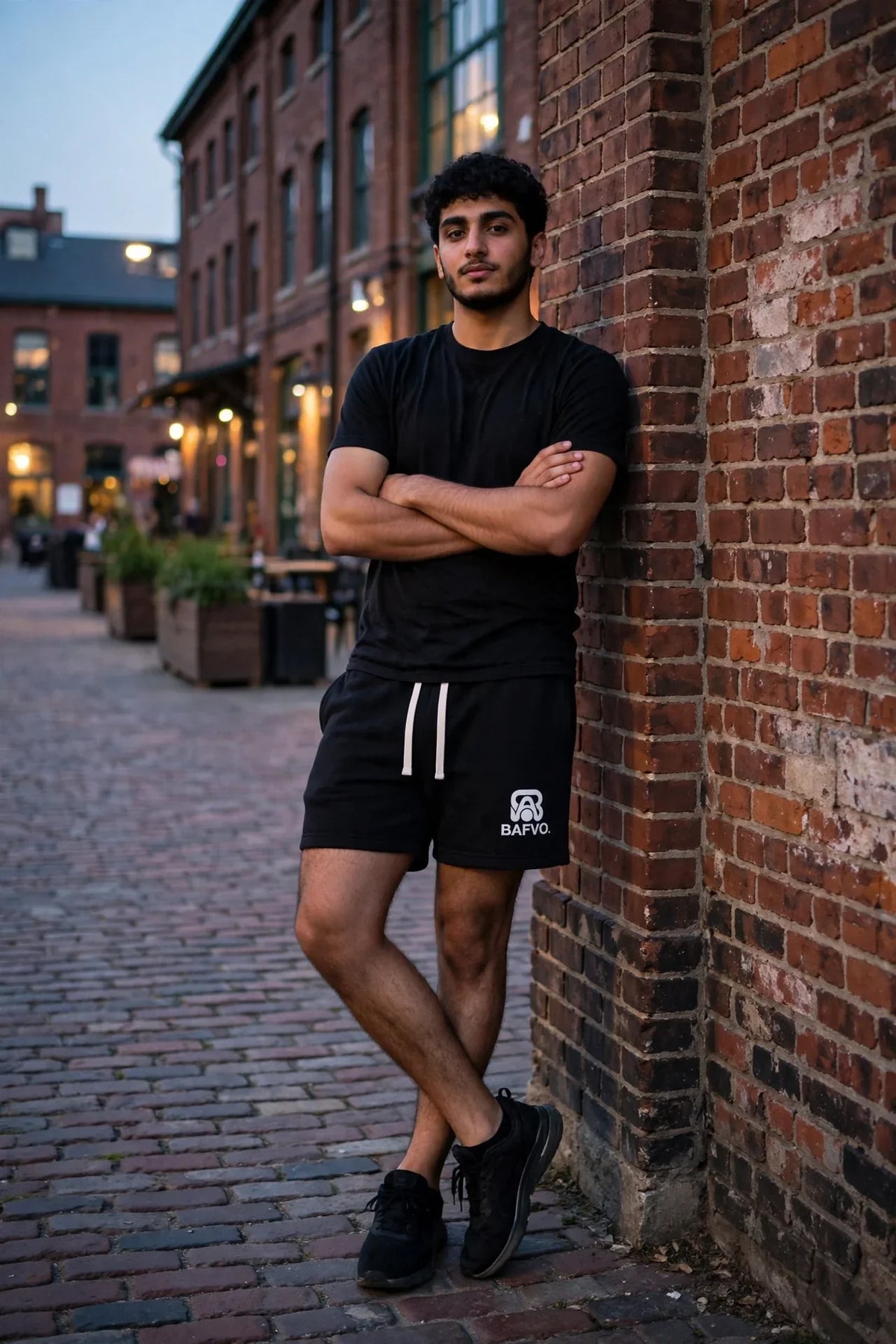 Man wearing black BAFVO fleece sweatshorts with white drawstrings and a black t-shirt, leaning against a brick wall in Toronto's Distillery District at dusk